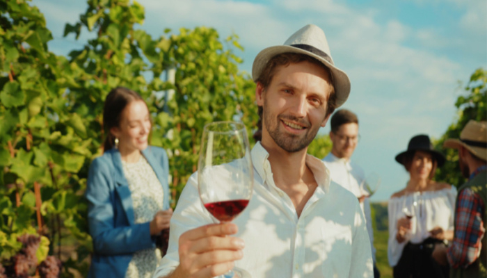 Happy young man smiling holding glass of wine on a wine tour experience with group of friends in vineyard